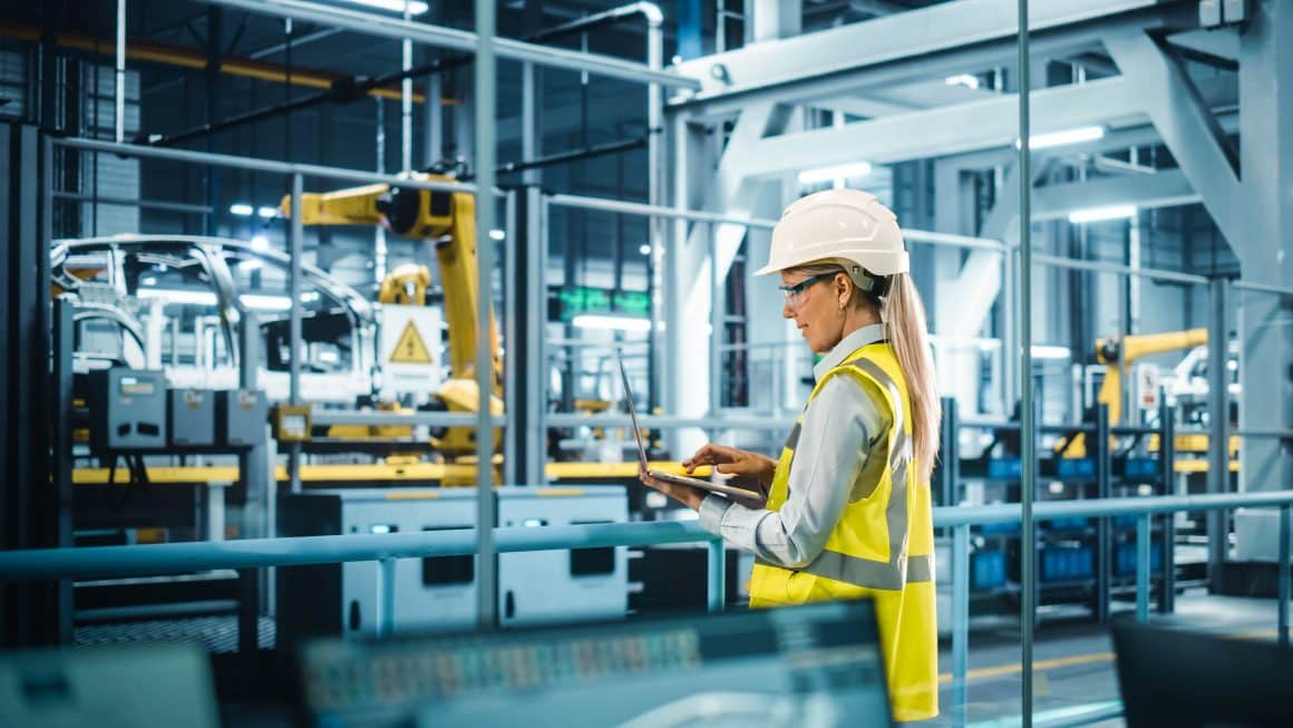 Car Factory: Female Automotive Engineer Wearing Hard Hat, Standing, Using Laptop. Monitoring, Control, Equipment Production. Automated Robot Arm Assembly Line Manufacturing Electric Vehicles.